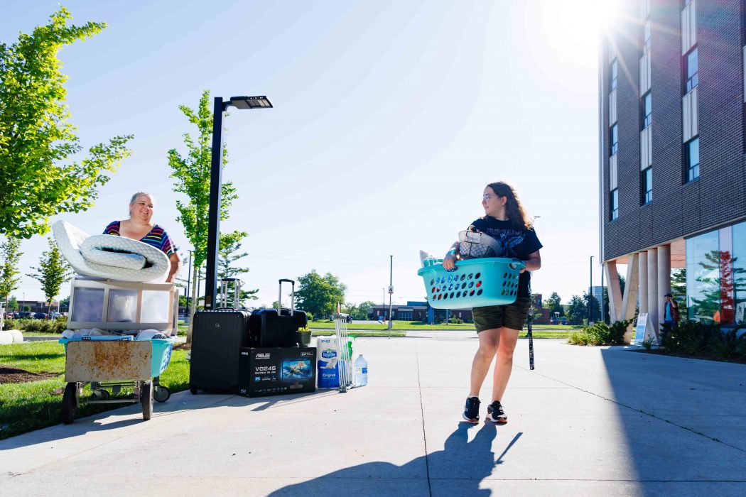 Two woman move household items across a cement concourse towards a building.