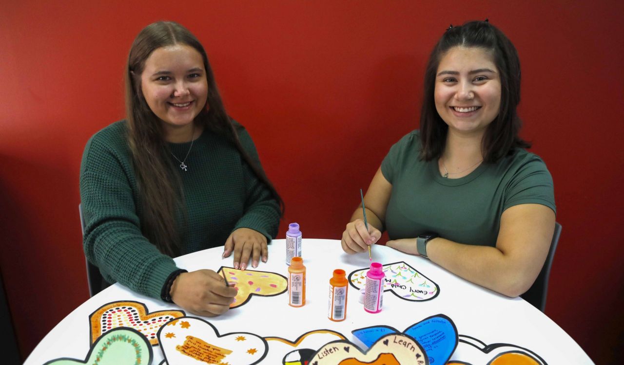 Evelyn Dilworth and Sophia Carnovale sit side-by-side at a table in front of a red wall and paint messages onto paper hearts.