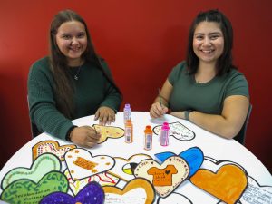 Evelyn Dilworth and Sophia Carnovale sit side-by-side at a table in front of a red wall and paint messages onto paper hearts.