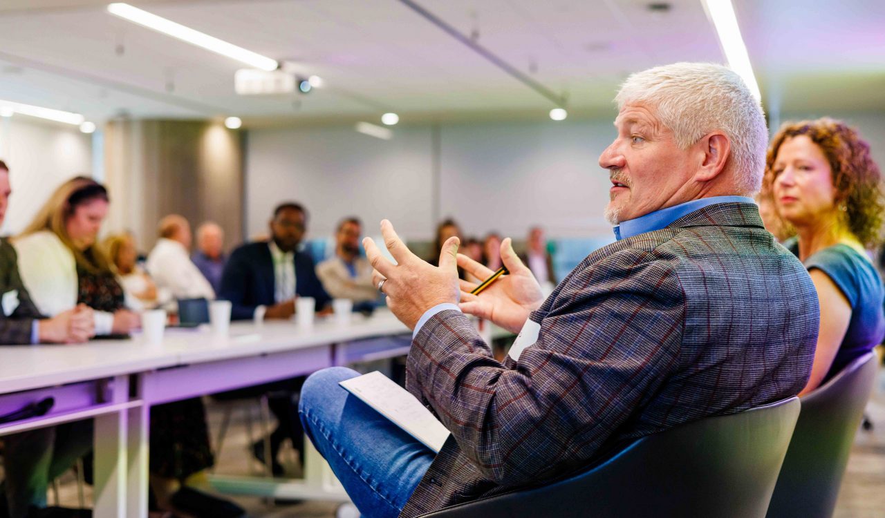 A man sits in front of a seated crowd while discussing a professional panel.