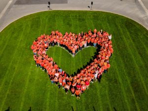 Dozens of people in orange shirts form a heart shape that can be seen from above. They are standing on green grass.