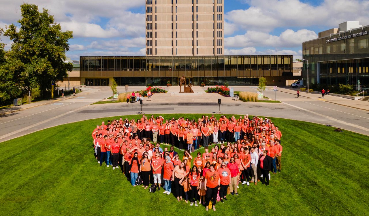 Dozens of people in orange shirts form a heart shape that can be seen from above. They are standing on green grass in front of Brock's Schmon Tower and Rankin Familiy Pavilion.