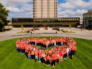 Dozens of people in orange shirts form a heart shape that can be seen from above. They are standing on green grass in front of Brock's Schmon Tower and Rankin Familiy Pavilion.