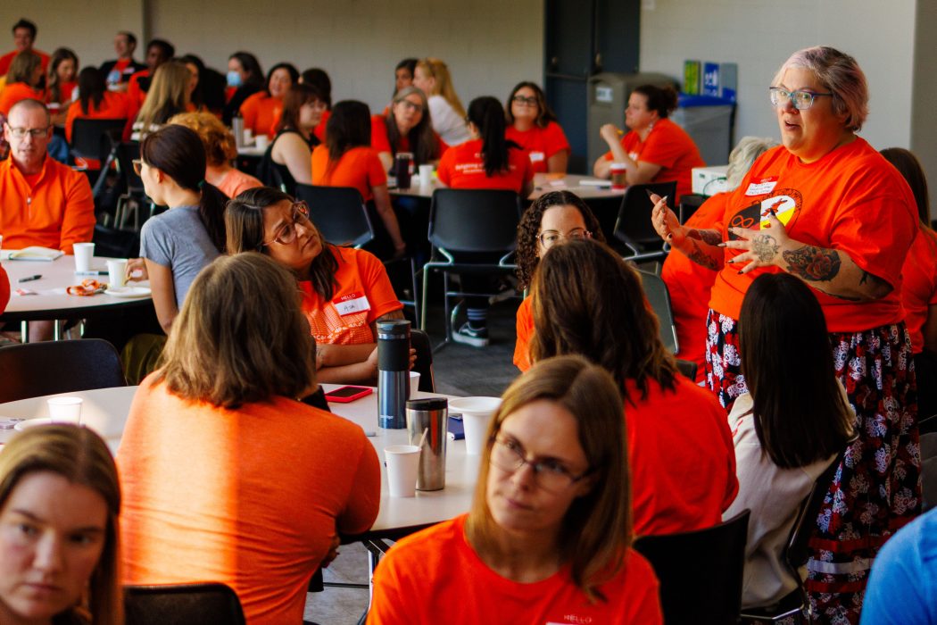 A woman wearing an orange shirt stands speaking to a large, seated group of people who are also wearing orange shirts to recognize the National Day for Truth and Reconciliation at Brock University’s Pond Inlet.