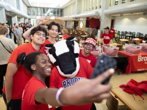 Four university students in red shirts take a selfie with a badger mascot in a cafeteria.