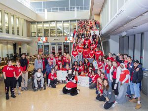 A large group of people in red shirts pose on and a round a staircase in a sunny university cafeteria.