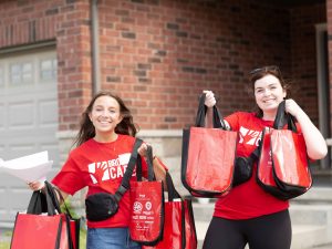 Two young women in red shirts hold up red gift bags outside a house.