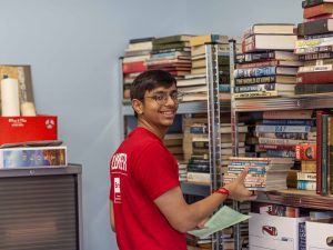 A young man in a red shirt sorts books on a shelving unit.