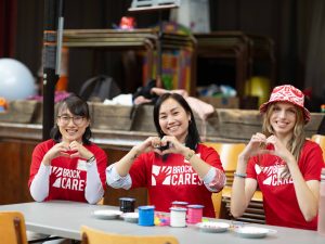 Three young women in red shirts pose, while making heart shapes with their hands, sitting at a table.