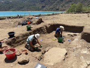 Two students dig at an archaeological site in the sand close to the ocean.