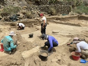 A group of students at work in a sandy area looking at the ground, studying archaeological finds.