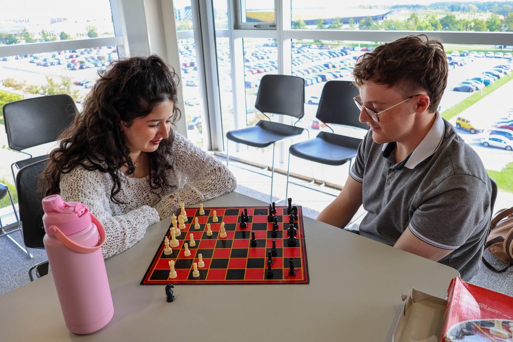 Two university students play a game of chess in front of a large window.