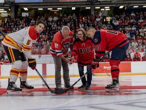 Four people stand on the ice of an arena dropping the puck to start a hockey game.