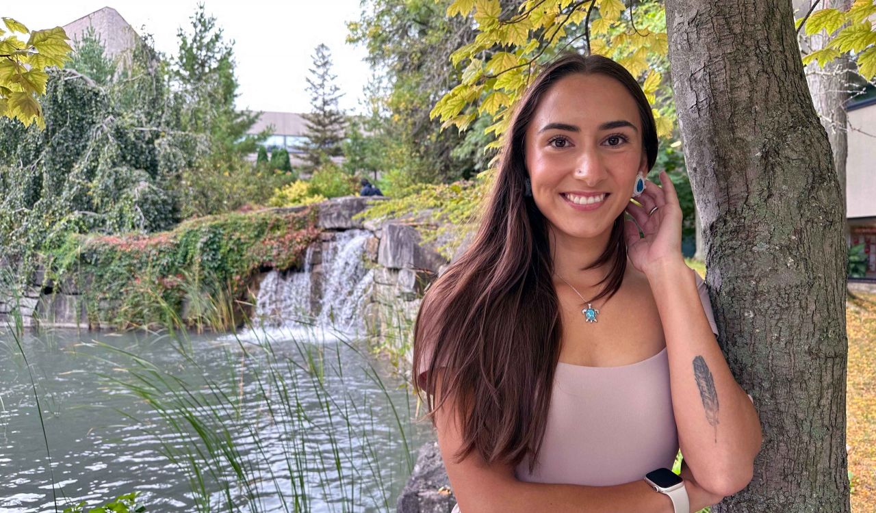 A young woman poses for a photo under a tree with a pond in the background.