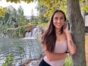 A young woman poses for a photo under a tree with a pond in the background.