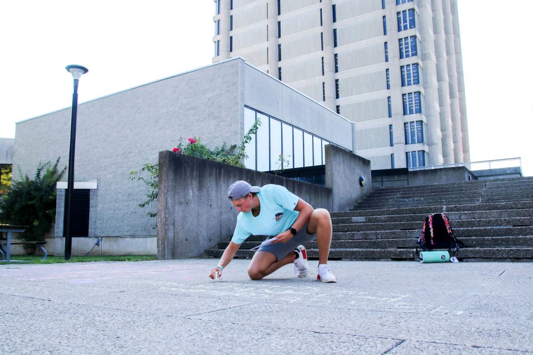 A university student writes on a sidewalk in chalk with a concrete tower in the background.