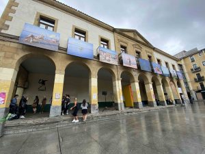 A yellow building with arches is adorned with artistic photographs in large format along the top of the building. Students stand under the building roof on an overcast day.