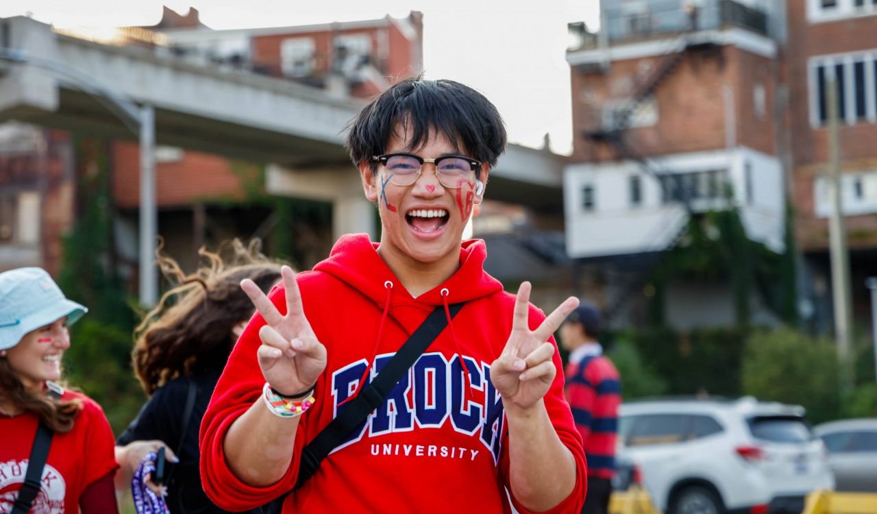 A man stands outdoors smiling giving the peace sign while wearing a red Brock University hoodie.