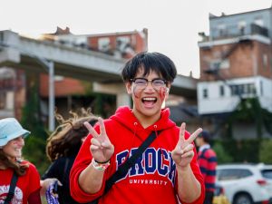 A man stands outdoors smiling giving the peace sign while wearing a red Brock University hoodie.