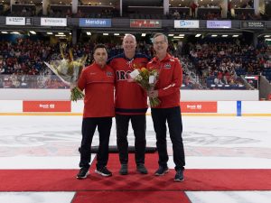 Three people stand together on the ice of an arena.