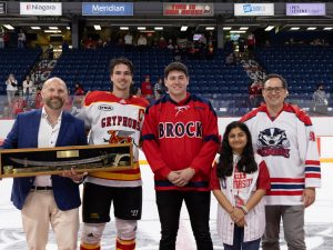 Five people stand on the ice of an arena, one holding a sword in a wooden case.