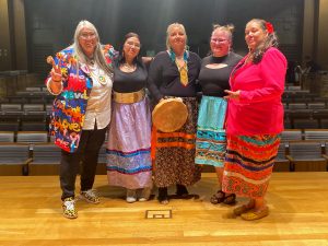 Five women, including four wearing ribbon skirts and one holding a drum, stand together on a stage.