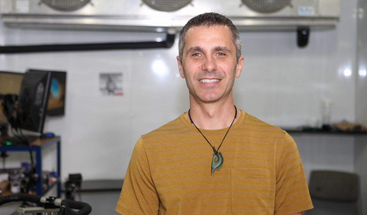 Head-and-shoulders photo of Canada Research Chair Toby Mündel smiling into the camera, with his lab machines blurred out in the background.