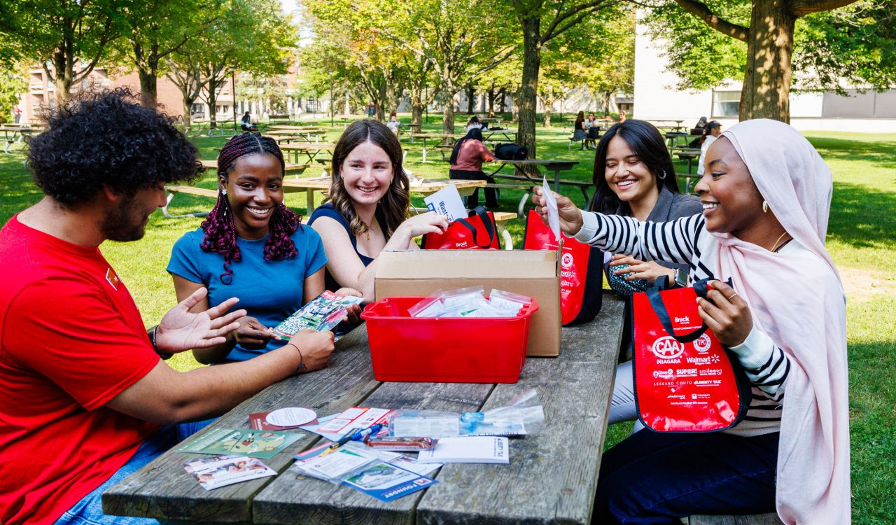 Five people sit at a picnic table putting together bags filled with brochures and other paper materials.