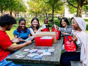 Five people sit at a picnic table putting together bags filled with brochures and other paper materials.