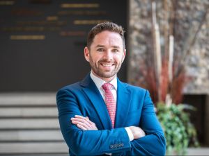 A man in a business suit poses for a photo in front of a building.