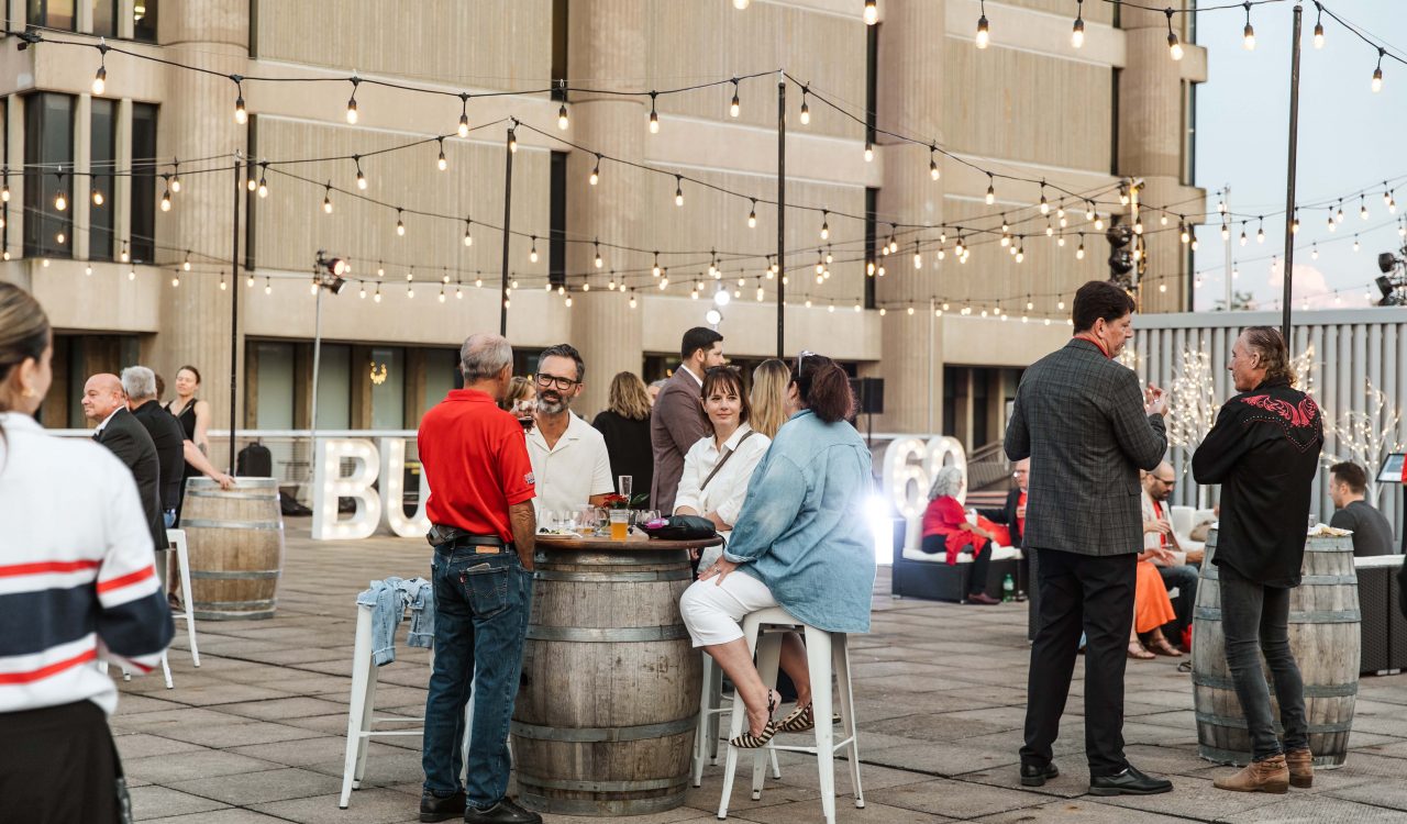 A group of people sit around a wine barrel table while at at outdoor event with string lights covering a patio area.