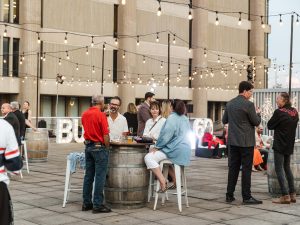 A group of people sit around a wine barrel table while at at outdoor event with string lights covering a patio area.