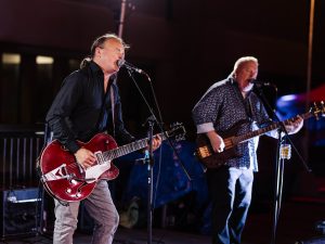 A band plays on stage with the singer performing into a microphone while playing a guitar as a bassist performs in the background.