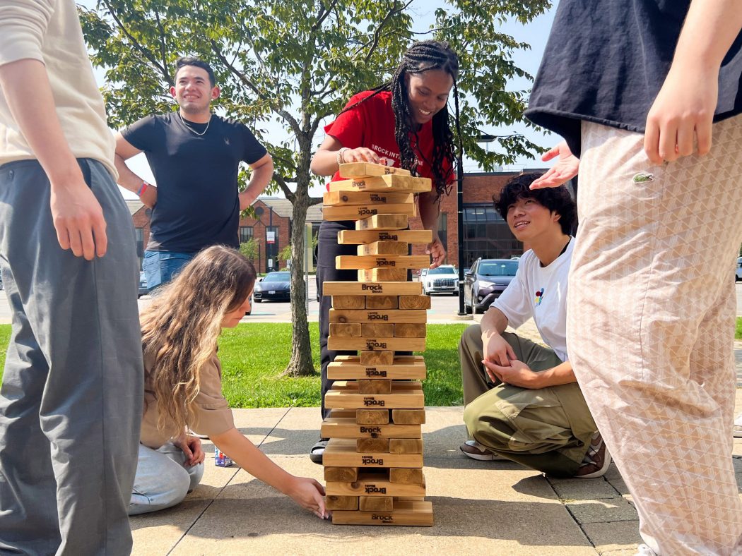 A group of international students stack giant Jenga blocks outside the Brock International Centre building on a sunny day.