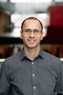 A man in glasses and a grey shirt poses for a headshot.