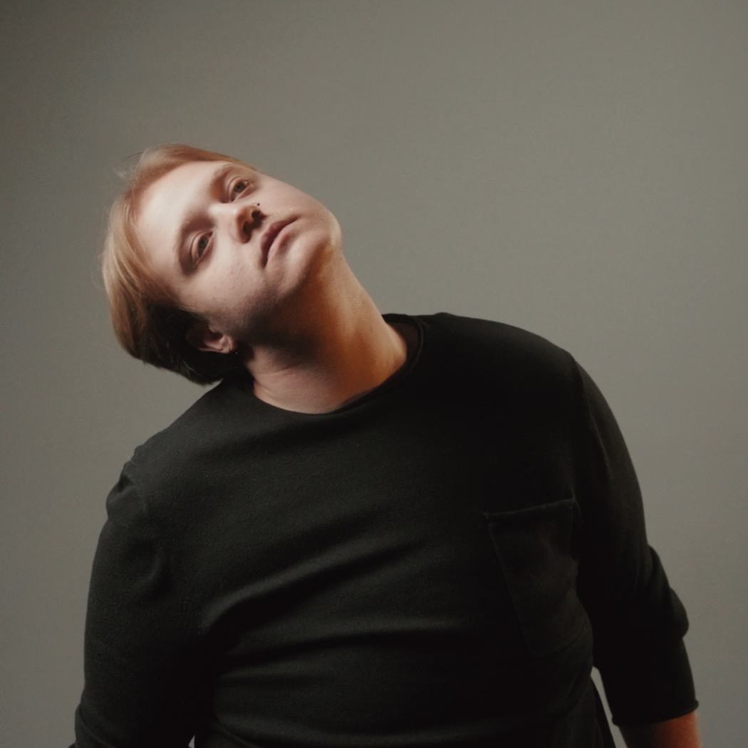 A portrait image of Vik Mudge shows them in a black shirt with their head to the side against a grey background.