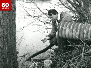 Then-Brock Biology Professor Michael Dickman squats next to, and leans his arm upon, a pipe jutting out of the ground covered by bushes and branches on the edge of the Welland River. He is examining something he is holding in his hand.