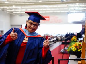 A graduate in a blue Convocation gown gives two thumbs-up while posing in front of an auditorium filled with people.