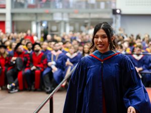 A graduate walks in front of a crowd of graduates and spectators at a Convocation ceremony.