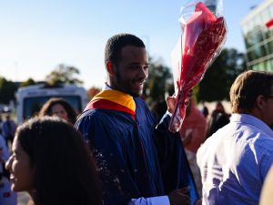 A graduate in a blue gown holds up a bouquet of flowers.