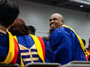 A graduate looks back and smiles while sitting with other graduates in a gymnasium.