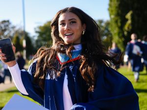 A graduate in a blue gown holds her cell phone while standing outside amongst other graduates.