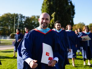 A graduate in a blue gown holds his degree while walking outside with other grads.