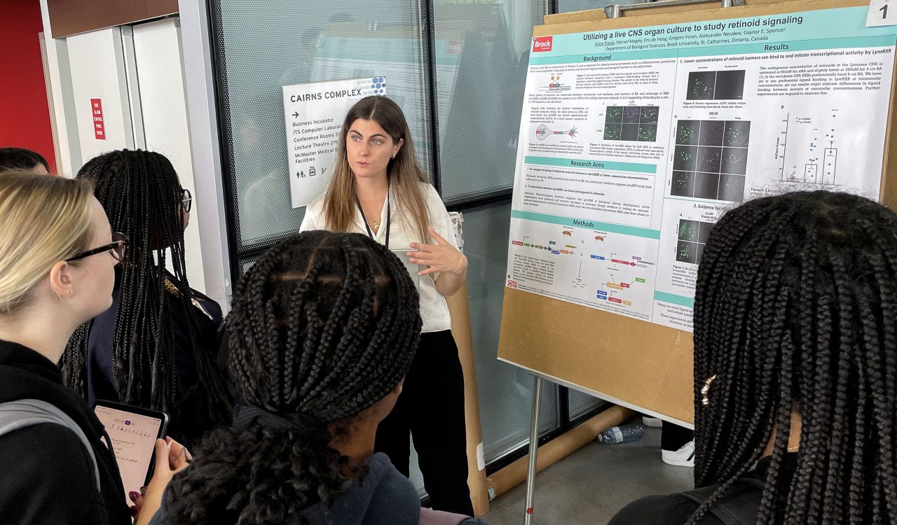 Alicia Piazza stands next to a poster that illustrates her PhD research as she speaks to a small crowd of people.