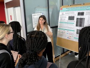 Alicia Piazza stands next to a poster that illustrates her PhD research as she speaks to a small crowd of people.