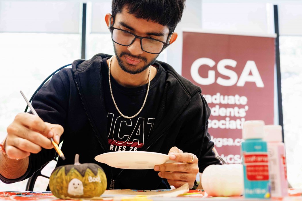 A man holds a paint brush over a green and black pumpkin with a large GSA sign in the background of a brightly lit room