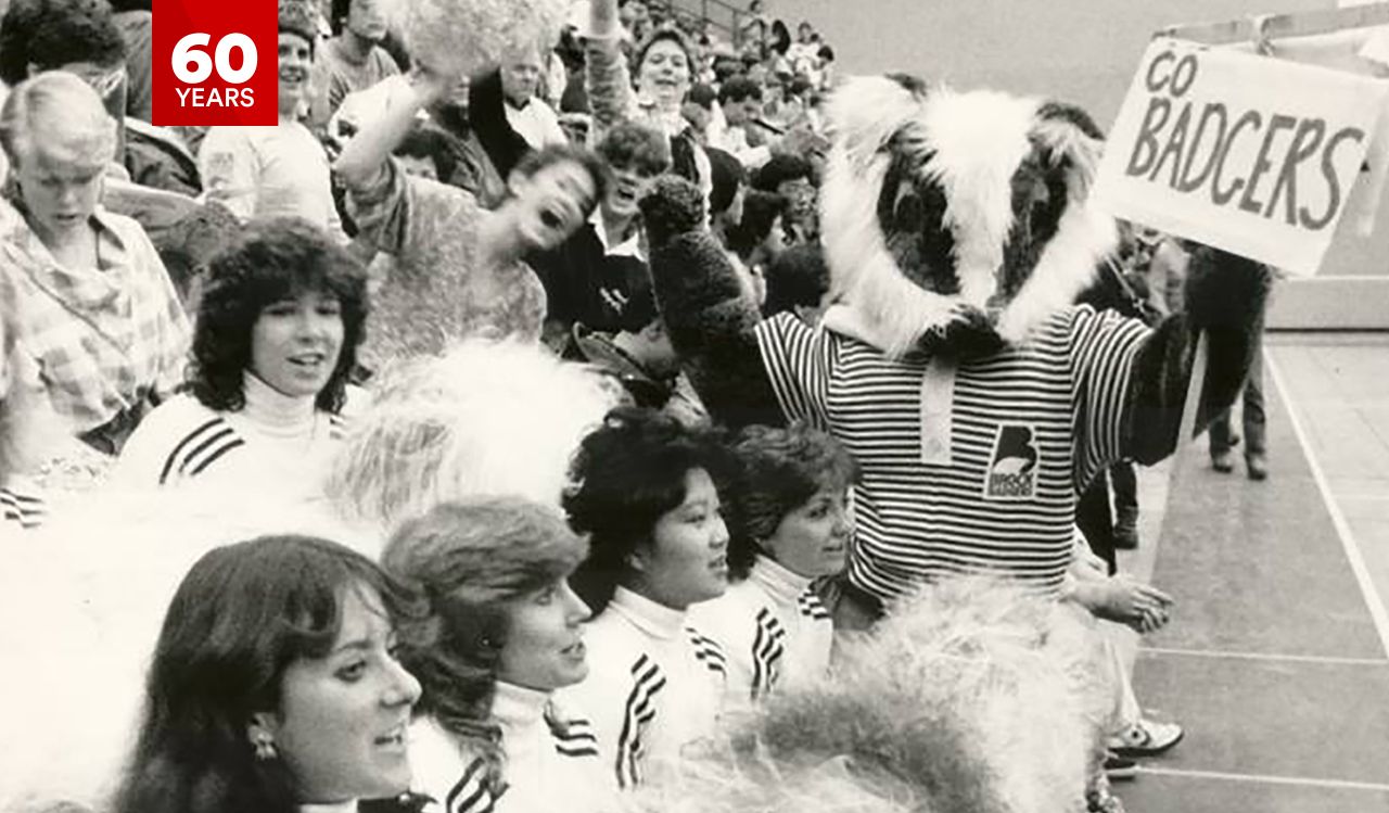 A furry mascot holds a Go Badgers! Sign amongst a crowd of fans and cheerleaders at a basketball game at Brock University.