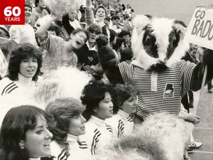 A furry mascot holds a Go Badgers! Sign amongst a crowd of fans and cheerleaders at a basketball game at Brock University.