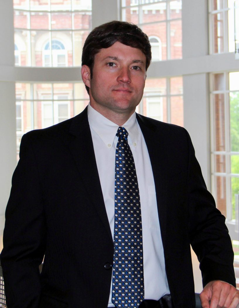 A man in a business suit poses for a photo in front of a window.