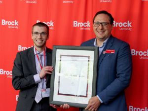 Two men in suits pose for a photo holding a framed certificate between them.